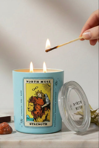 Candle with tarot card design labeled 'Strength' being lit with a match, on a neutral background.
